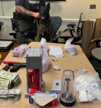 Table covered with seized items including bags of fentanyl, cash, a firearm, drug packaging equipment, and a bullet press, with a sheriff’s K9 and officer standing behind.