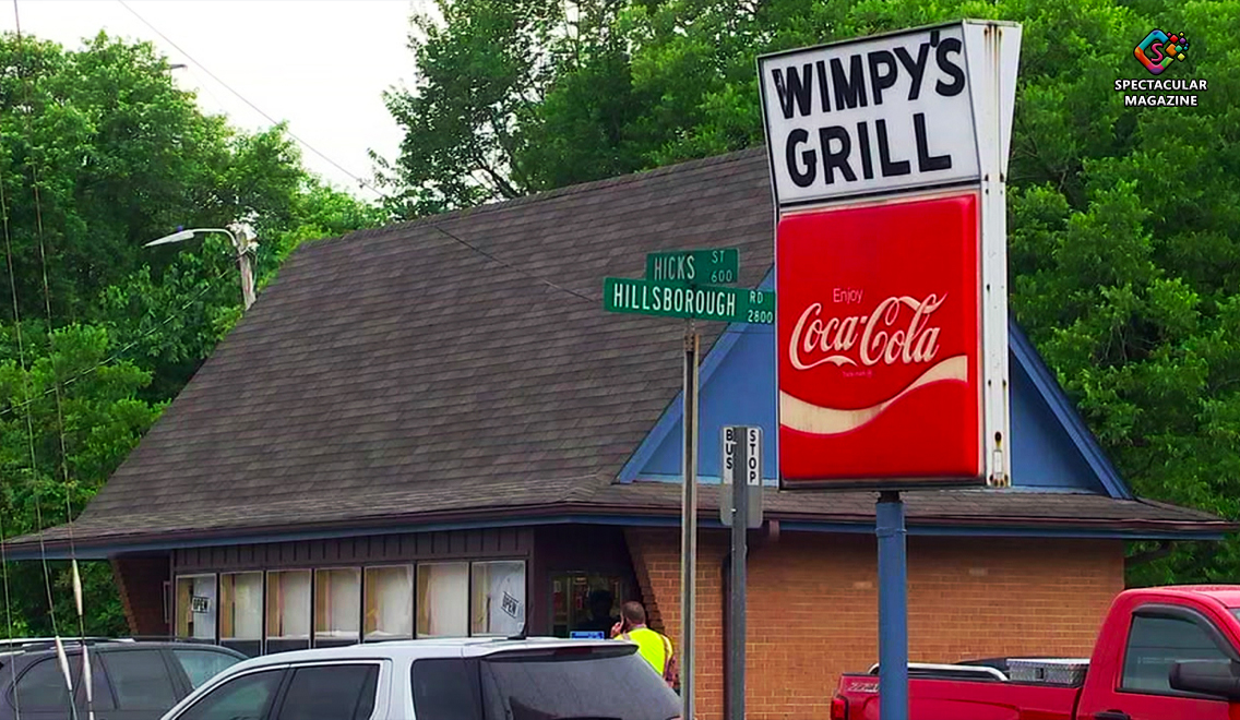 Exterior of Wimpy’s Grill in Durham, North Carolina, showing its iconic A-frame building, red Coca-Cola sign, and street signs for Hillsborough Road and Hicks Street.