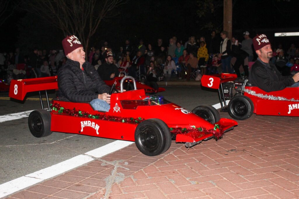 Red Shriners Fire Brigade go-karts driven in a nighttime parade, decorated with holiday lights and watched by a crowd along the street.
